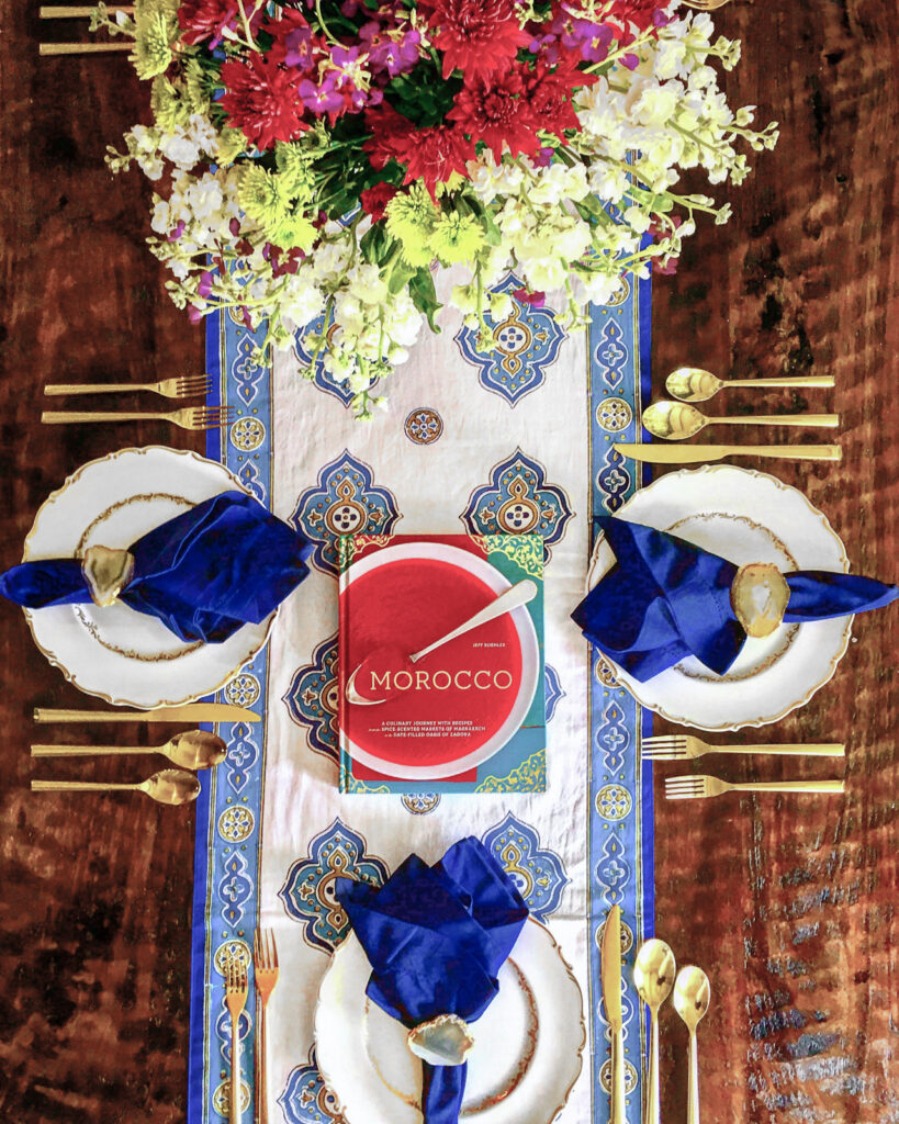 Moroccan blue and white table runner with red floral centerpiece, gold silverware, blue napkins, and a Morocco cookbook on the table runner