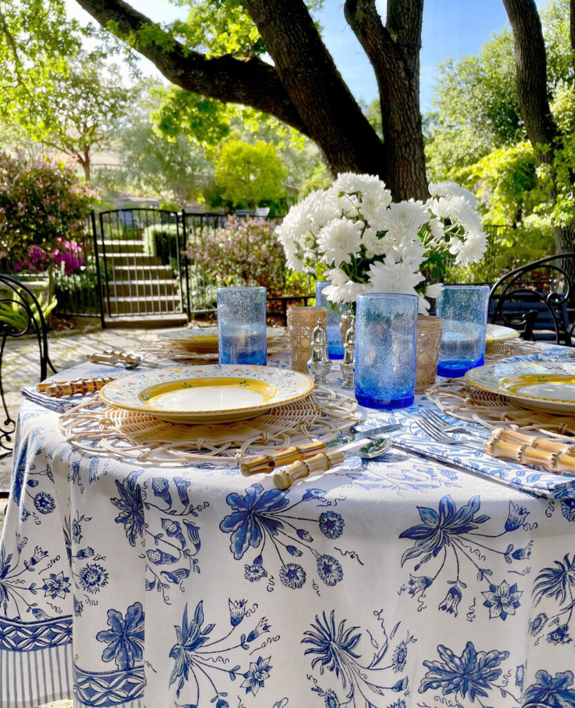 garden table outside on a sunny day with blue and white tablecloth, blue glasses, and flowers