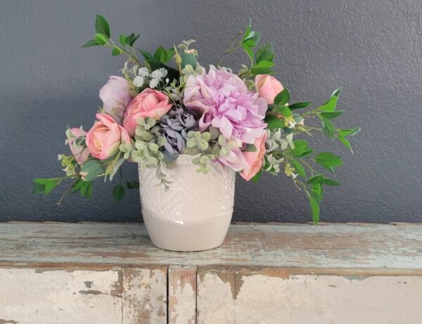 A centerpiece with ranunculus, peonies, roses, and decorative branches in a white vase on a white, distressed wood surface