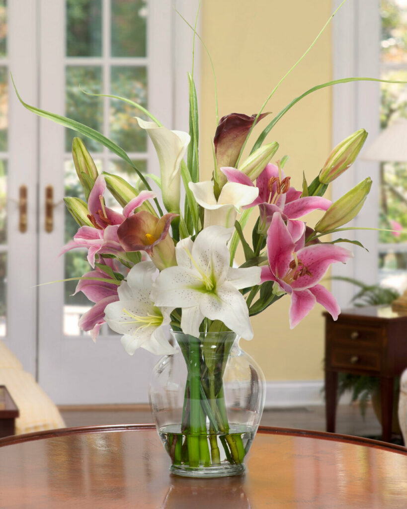 A floral arrangement  in a clear vase of white and pink lilies with leaves, buds, and blooms on a brown table