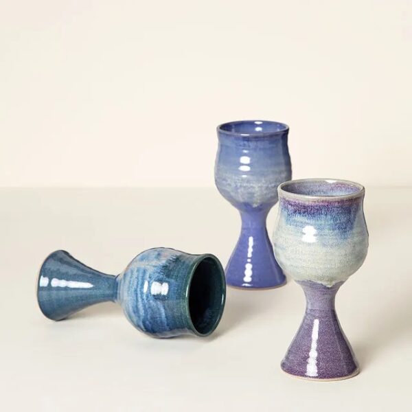 Three glazed and shiny blue and purple gradient goblets, two upright and one on its side against a deep cream background