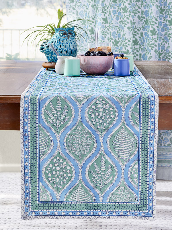A blue and green table runner inspired by woodland ferns with matching turquoise, lavender, and pink pastel cups and vase