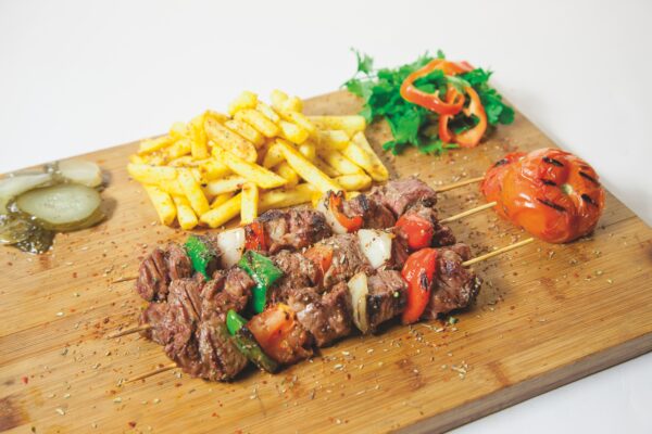 Photograph of barbecue and vegetable skewers, french fries, salad, and pickles on a wooden board