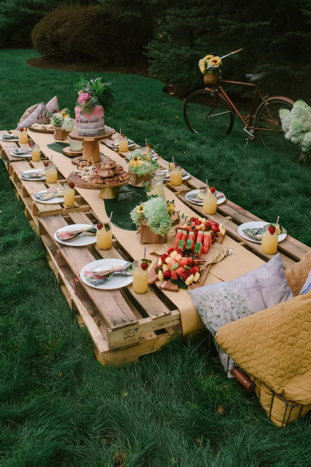 Photo of pallet table with tropical drinks, fruits, and a cake on the table runner as an engagement party idea
